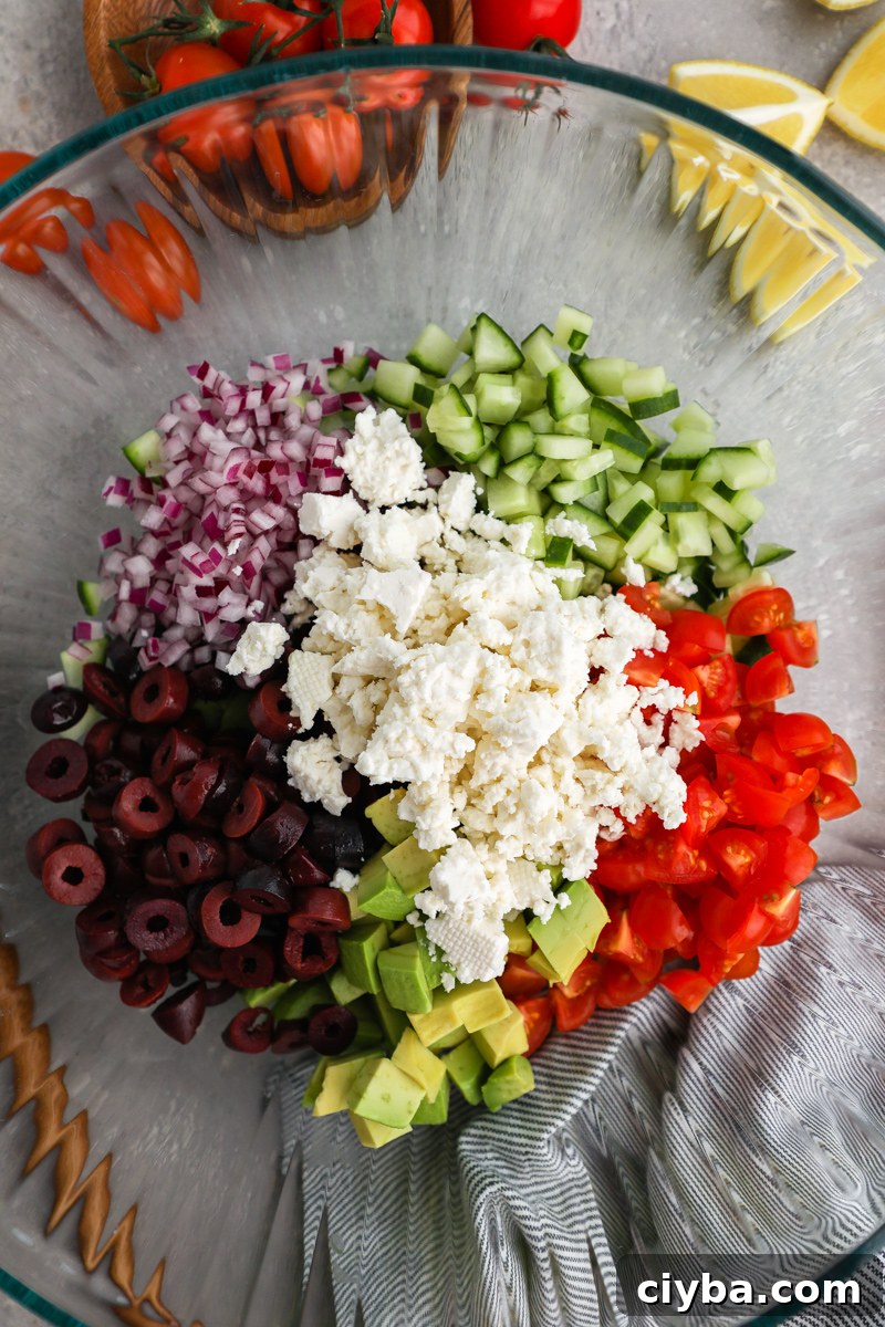 Mediterranean Avocado and Feta Salsa Recipe 4 A large clear mixing bowl filled with chopped cucumber, tomatoes, avocado, Kalamata olives, red onion and crumbled feta cheese sitting next to fresh lemon wedges