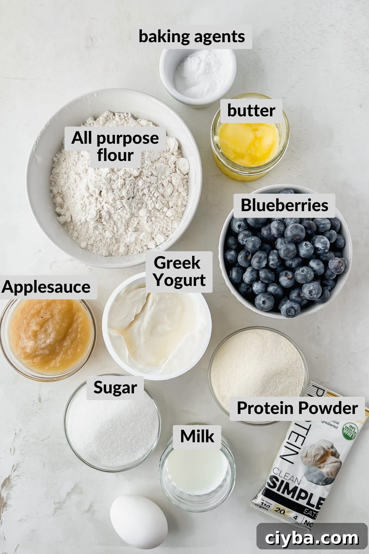 ingredients for blueberry protein muffins in individual bowls arranged on white background.