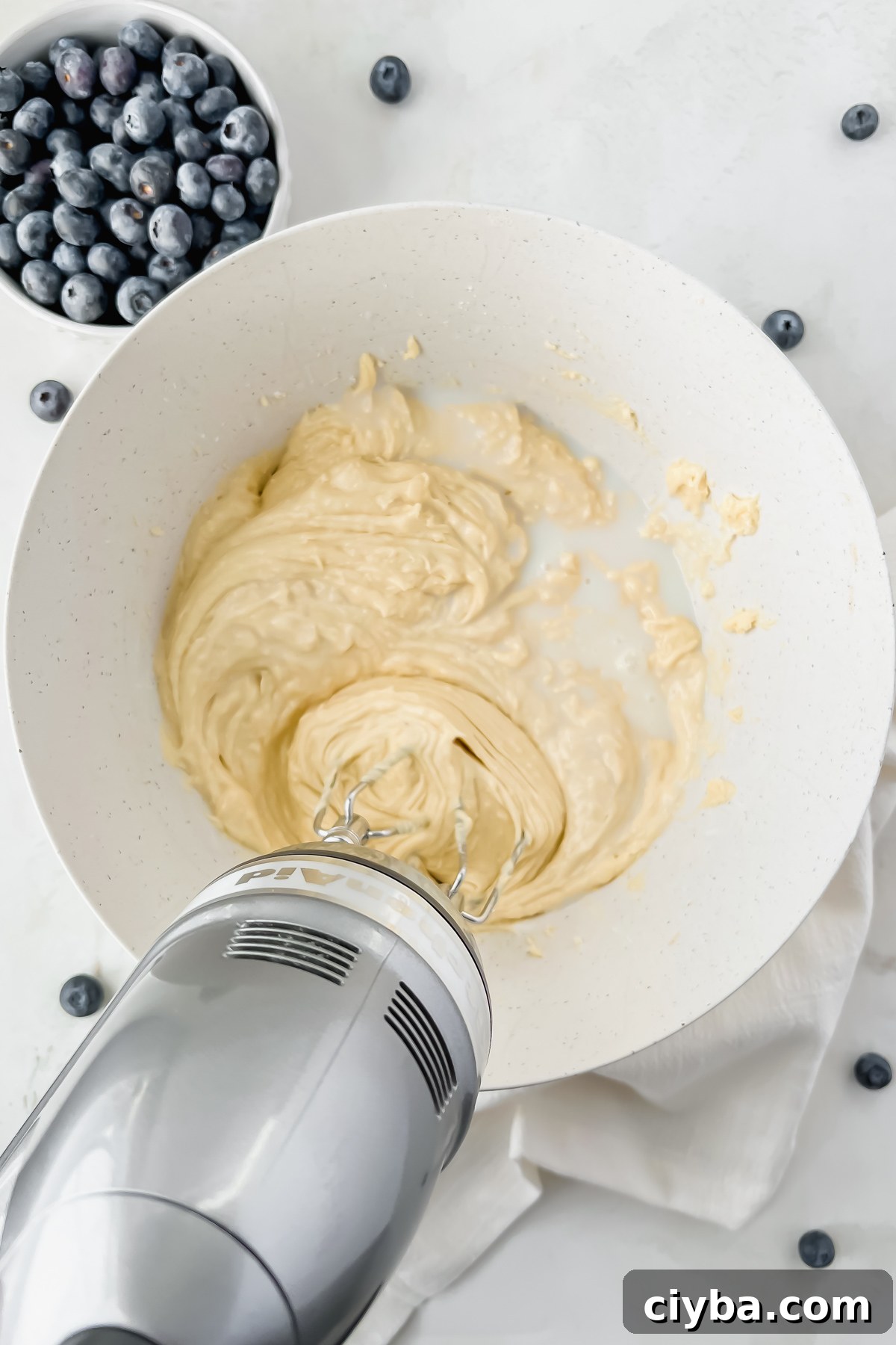 electric hand mixer mixing blueberry protein muffin batter in white mixing bowl.