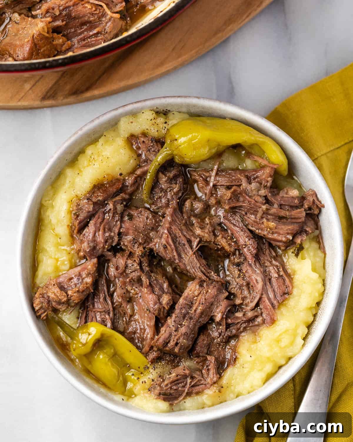 A bowl of mashed potatoes topped with Mississippi pot roast and peperoncini sitting on a table.