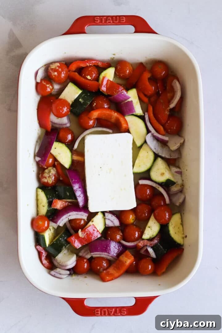 Raw vegetables and a block of feta in a large baking dish tossed in oil
