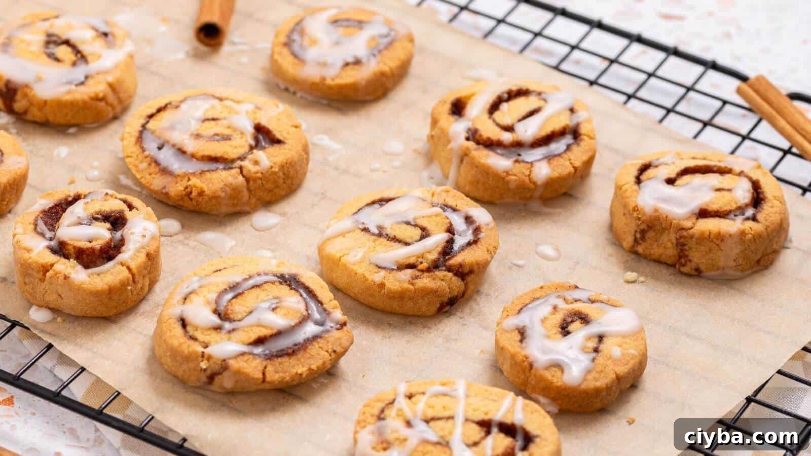 Cinnamon roll cookies with icing are placed on a sheet of parchment paper on a cooling rack. Two cinnamon sticks are visible beside them.