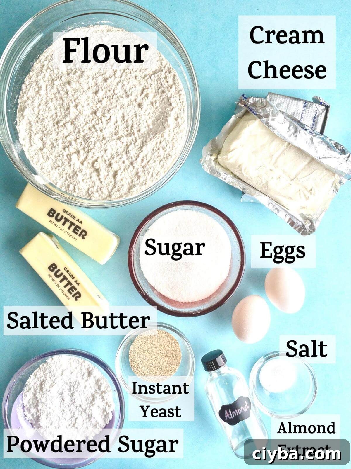 An overhead shot of cream cheese danish ingredients with the following text: flour, sugar, salted butter, eggs, salt, instant yeast, cream cheese, powdered sugar, almond extract.
