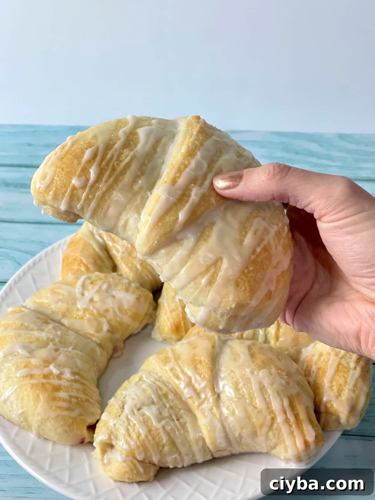 A picture with a hand holding a large danish with cream cheese filling, with a plate of danish drizzled with a powdered sugar glaze in the background.
