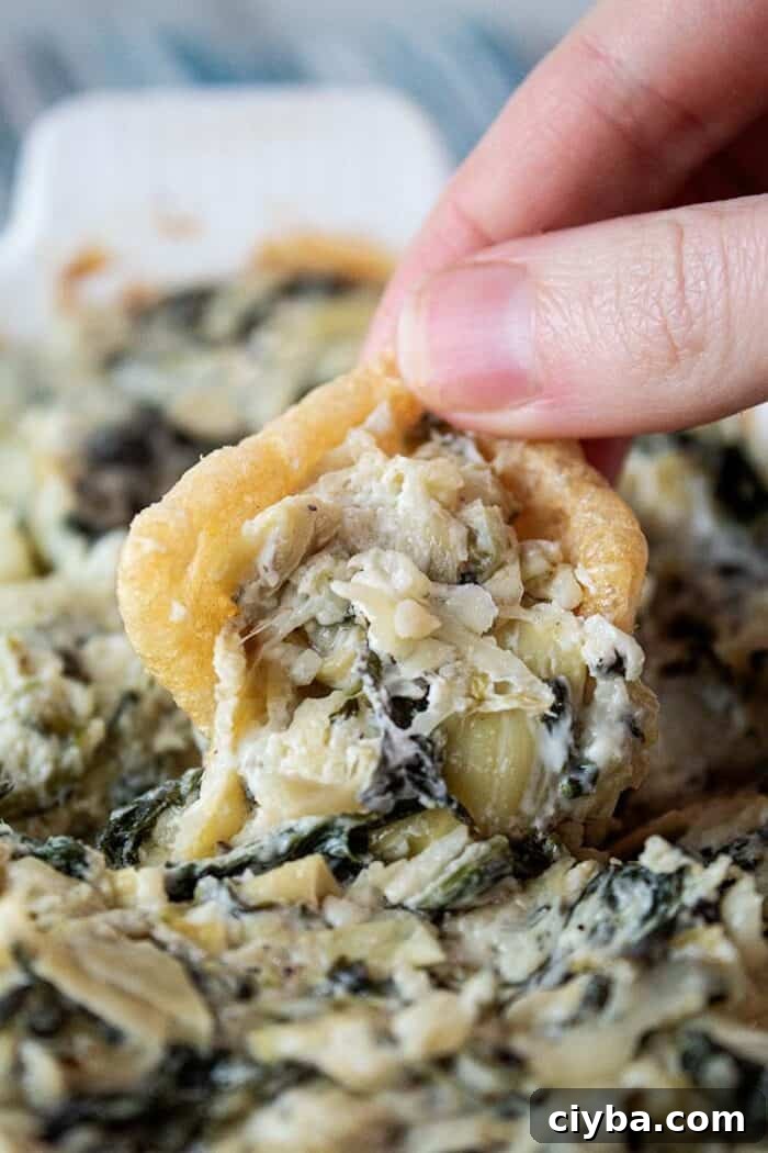 Keto Super Bowl Party Menu: Low-Carb Game Day Recipes 6 sideview of a person's hand scooping a serving of keto spinach artichoke dip with a pork rind