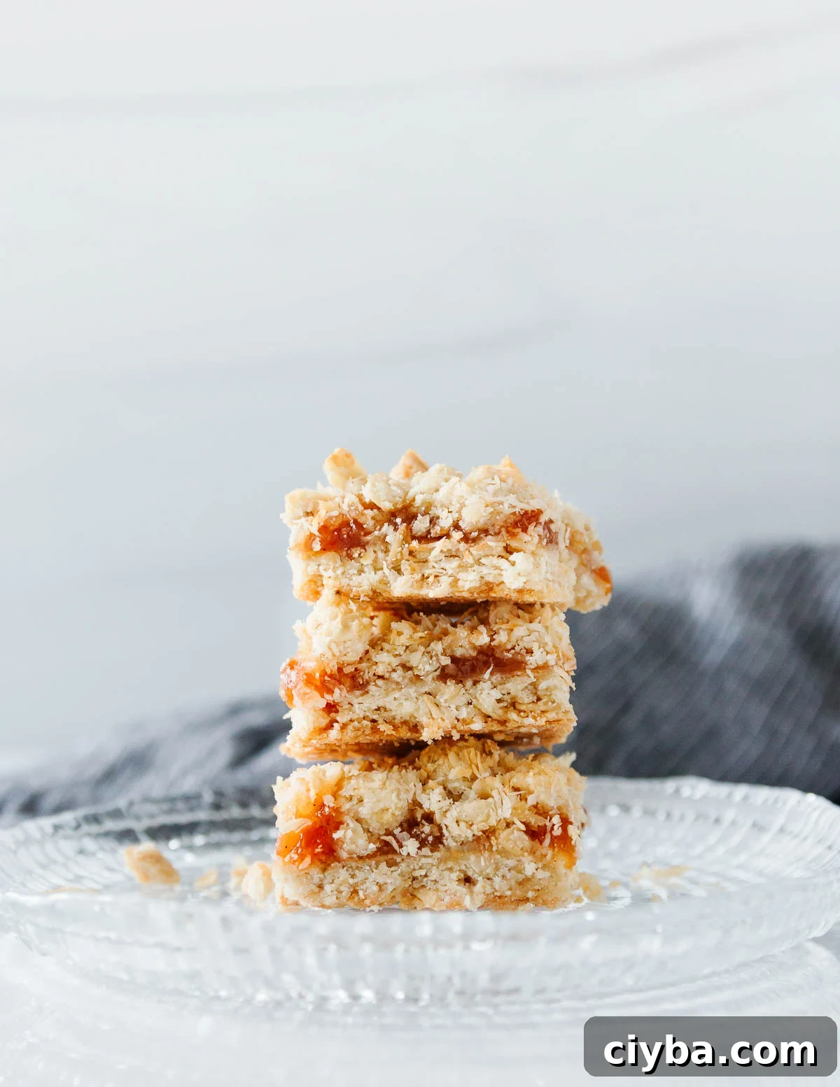 Coconut Peach Crumb Bars Recipe 2 Three stacked coconut peach crumb bars on a cut glass plate with a dark grey napkin in the background.