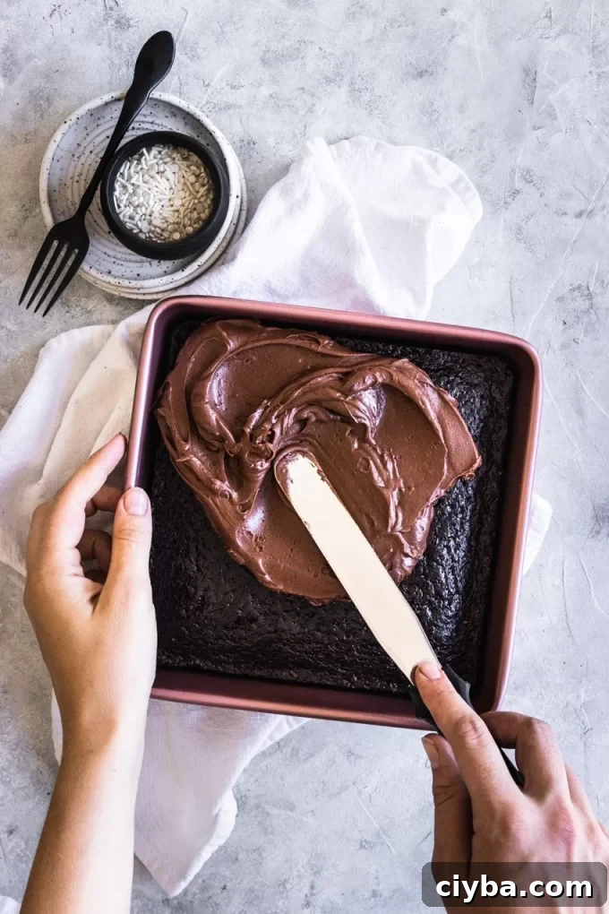 Aerial photo of a hand spreading chocolate frosting onto a sourdough chocolate cake in a pan.