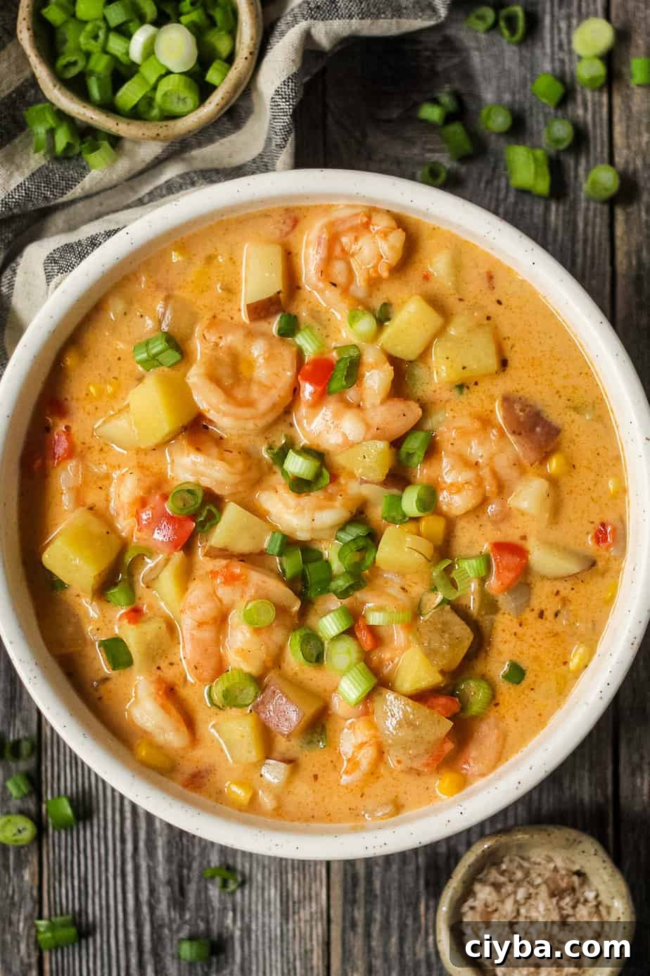 Creamy Cajun Shrimp and Corn Chowder Recipe 2 overhead shot of creamy cajun shrimp and corn chowder in a white bowl on a wooden surface. Bowl of salt and chopped green onions in top left and bottom right of shot.
