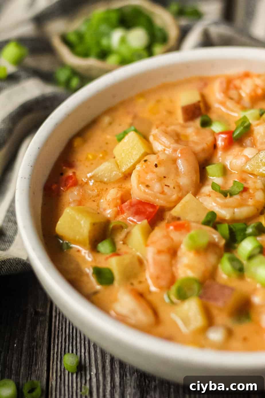 Creamy Cajun Shrimp and Corn Chowder Recipe 3 side angle shot of creamy cajun shrimp and corn chowder in a white bowl on a wooden surface. Bowl of salt and chopped green onions in top left and bottom right of shot.