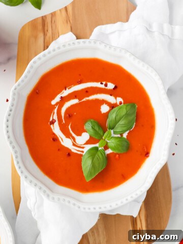 A white bowl filled with a smooth red soup. The soup is garnished with a drizzle of white coconut milk, fresh basil leaves, and crushed red pepper flakes.