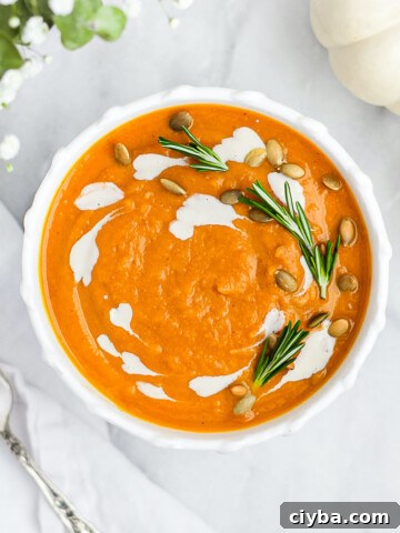 A white bowl with orange soup inside. There is white vegan crema, fresh rosemary, and pumpkin seeds garnishing the top.