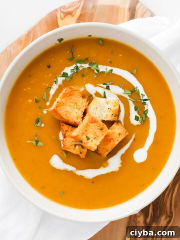 A white bowl filled with orange soup. The soup is garnished with a drizzle of white coconut milk, croutons, and chopped green herbs.