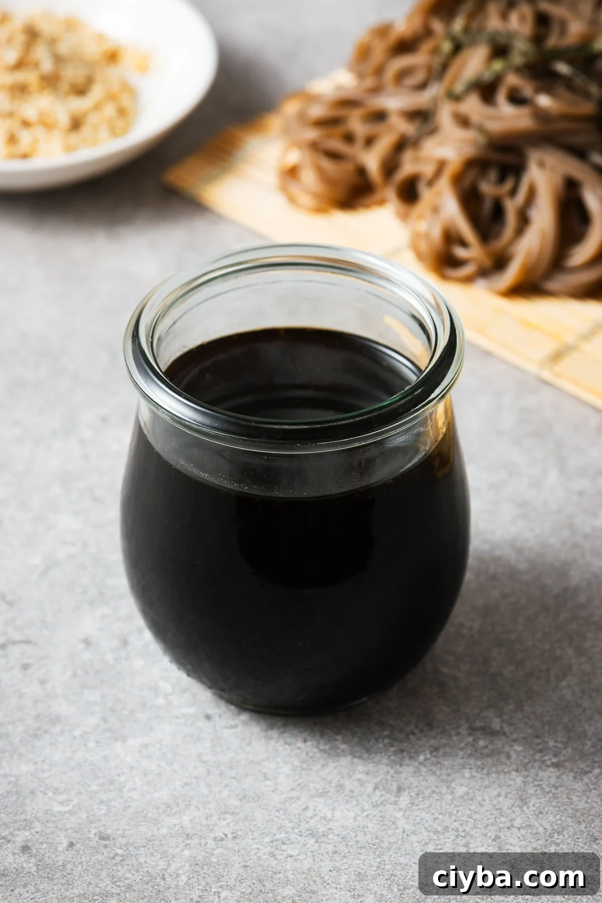 A weck jar with mentsuyu  and soba noodles and sesame seeds in the background.