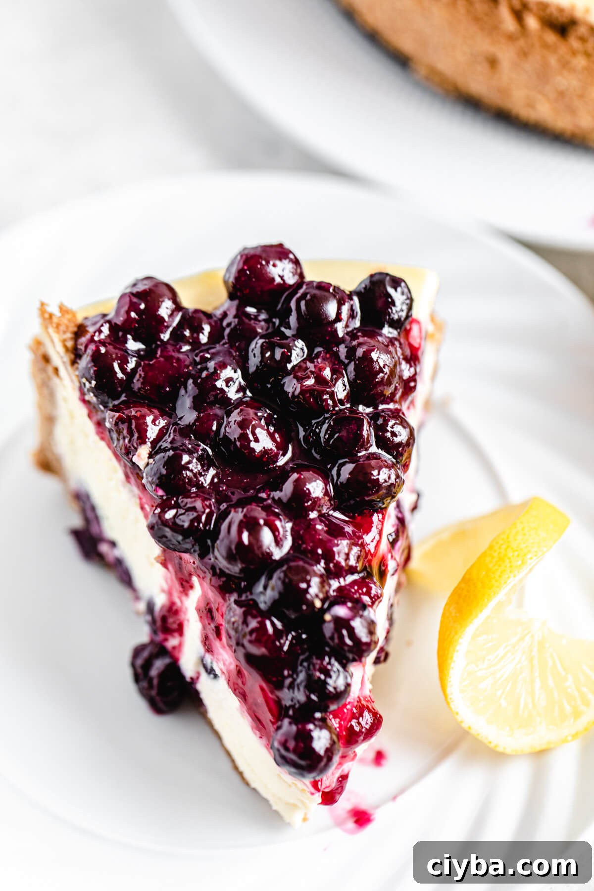 cheesecake slice topped with blueberry sauce on a white plate with lemon twist beside it