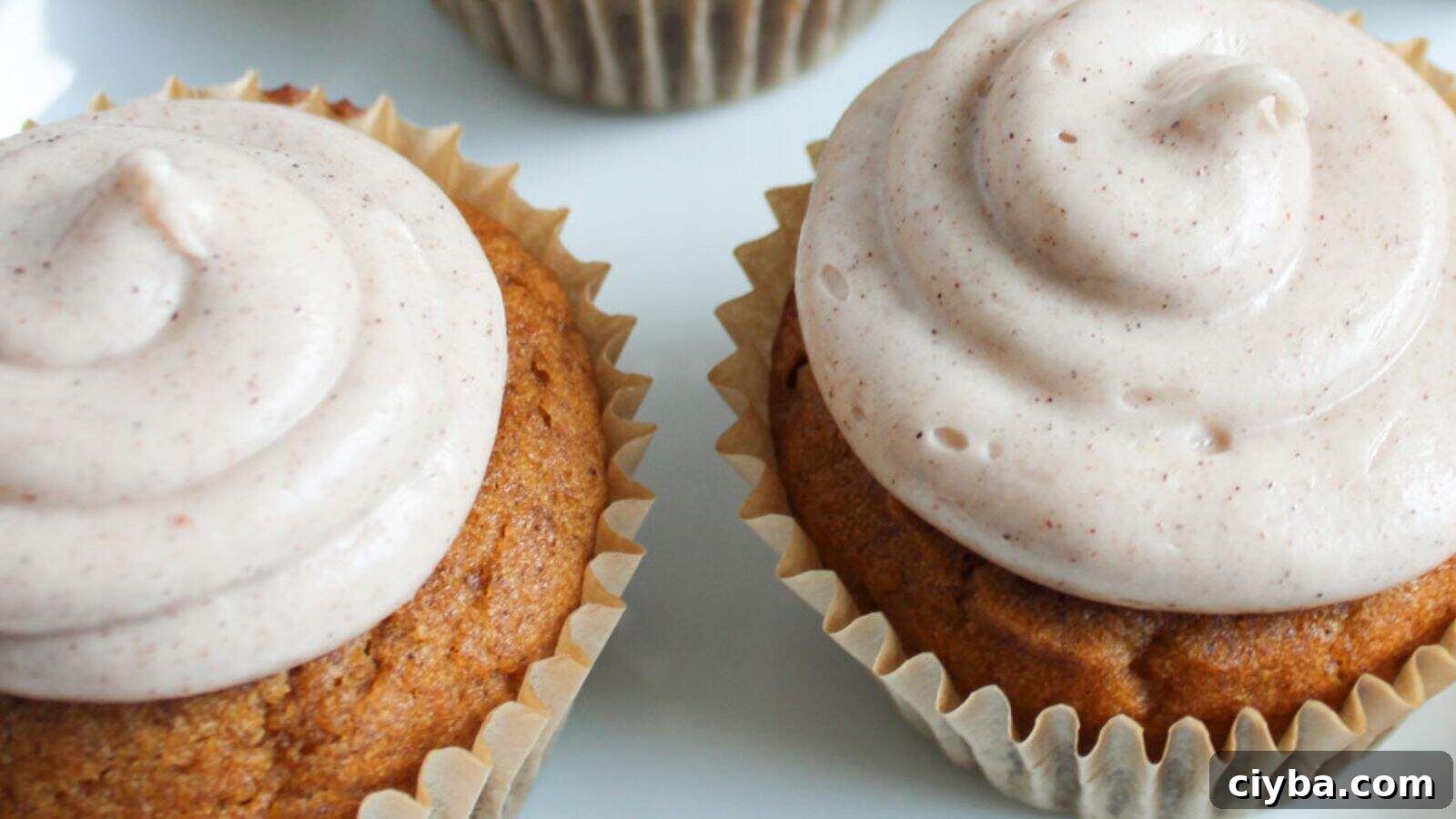 Two cupcakes with light brown frosting are shown on a white surface, each in a beige paper liner.