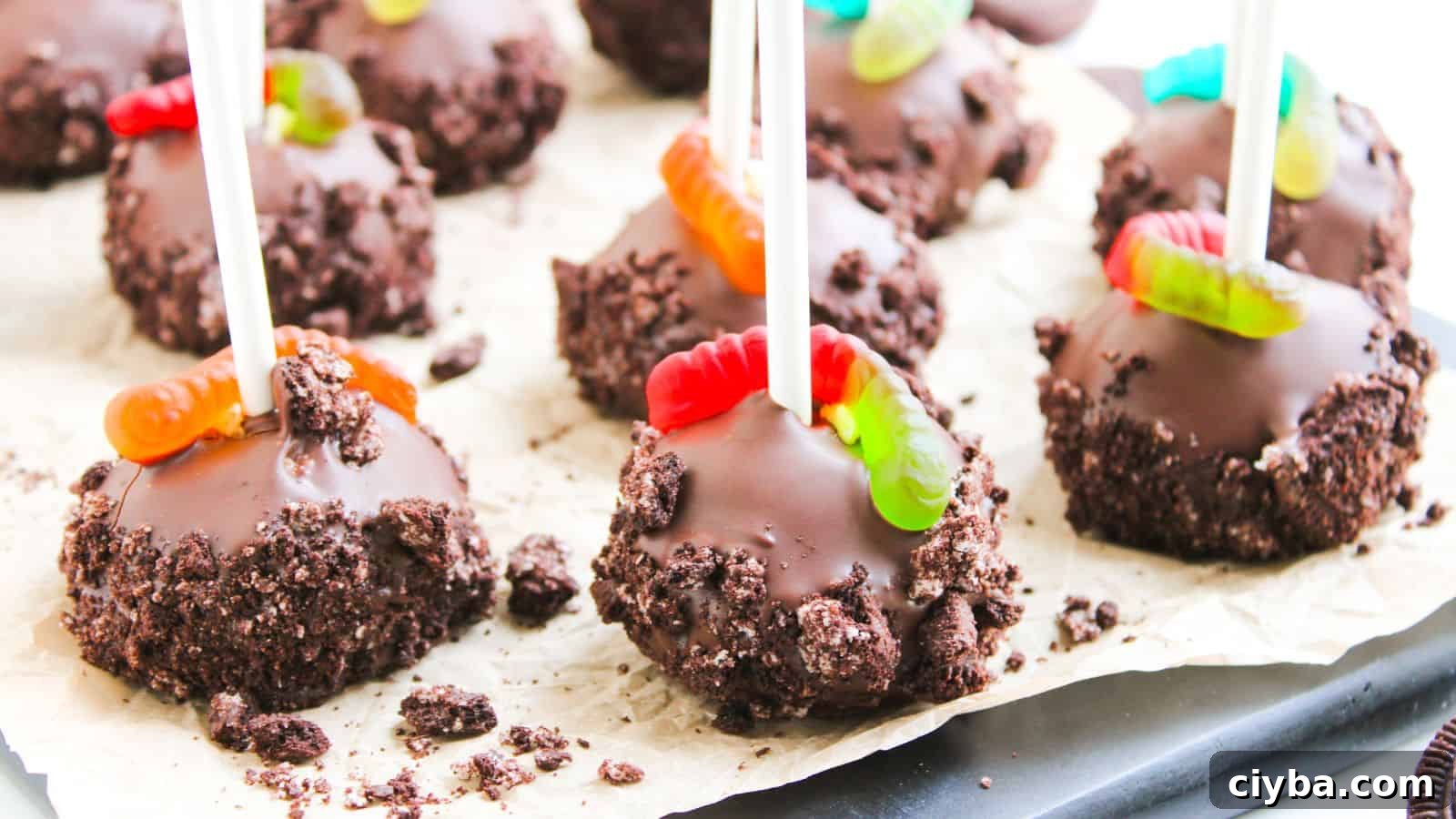 Chocolate cake pops covered in crushed cookies, each with a gummy worm on top, displayed on parchment paper.