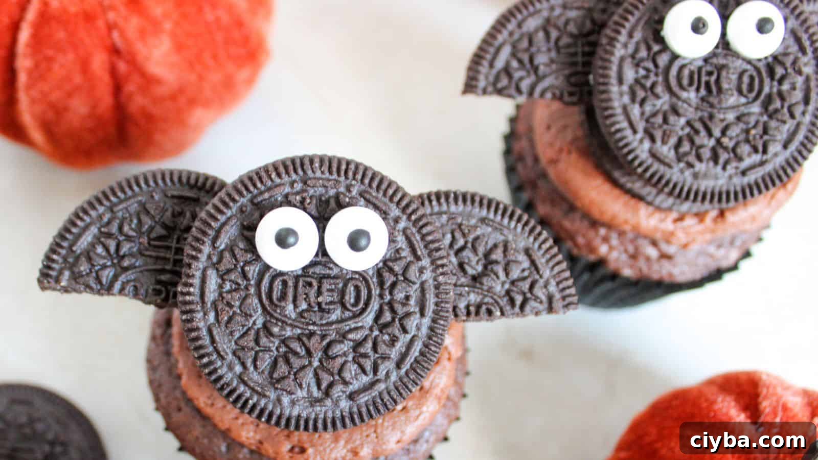 Chocolate cupcakes decorated with Oreo cookies resembling bats, complete with candy eyes. Orange pumpkins are visible in the background.