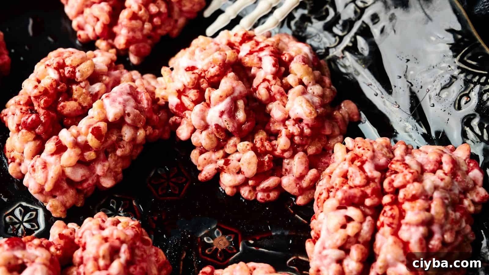 Clusters of pink and red puffed rice cereal treats are arranged on a dark, patterned plate next to a white skeleton hand decoration.