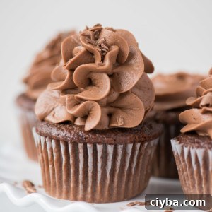 Moist Chocolate Cupcakes Recipe - Classic Homemade 5 A close-up of easy chocolate cupcakes with swirled chocolate frosting and chocolate shavings on top, placed on a white plate with more cupcakes in the background.
