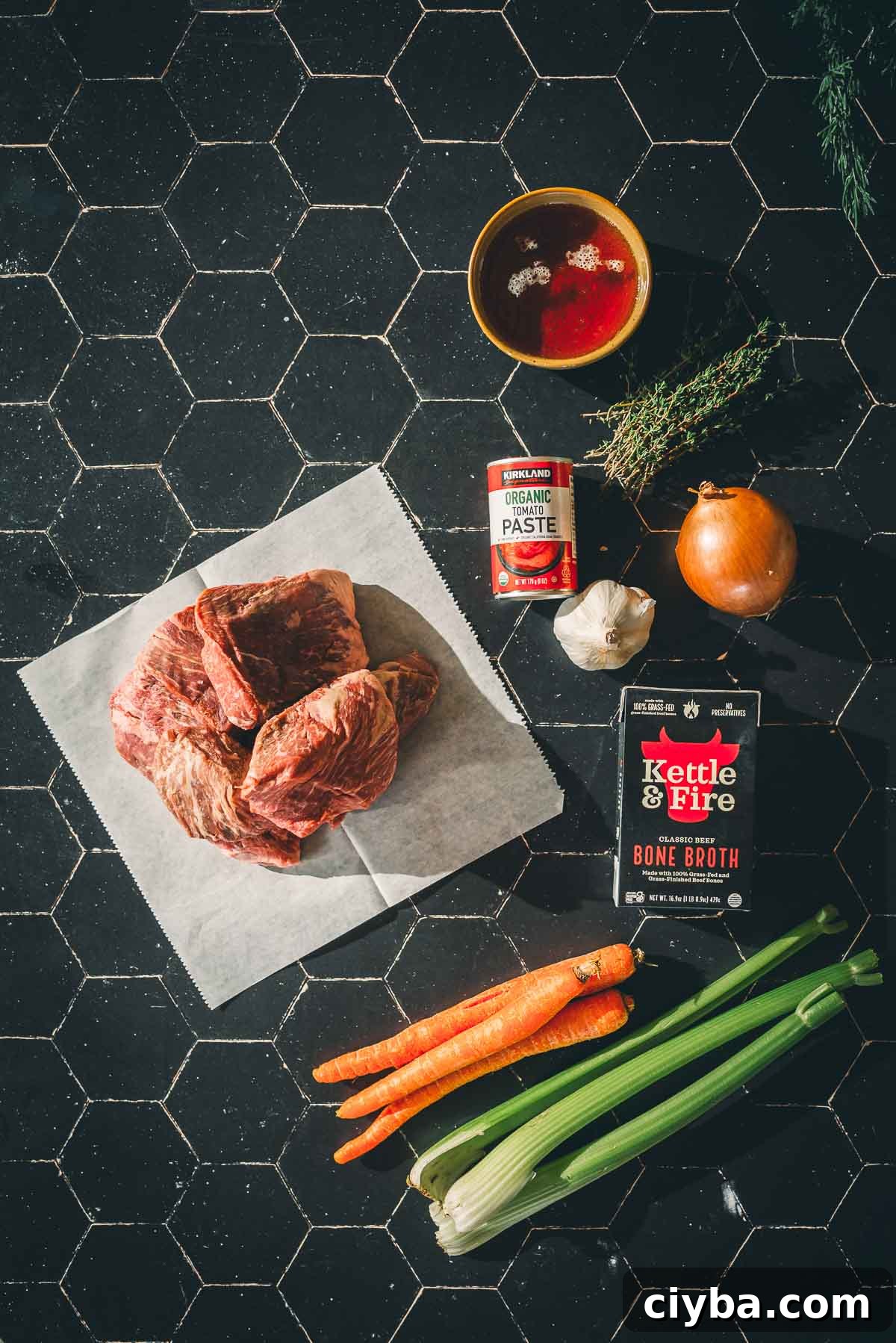 Ingredients for sous vide short ribs on a dark tile surface: raw beef on paper, tomato paste, bone broth, garlic, onion, carrots, celery, thyme, and a bowl of red liquid.