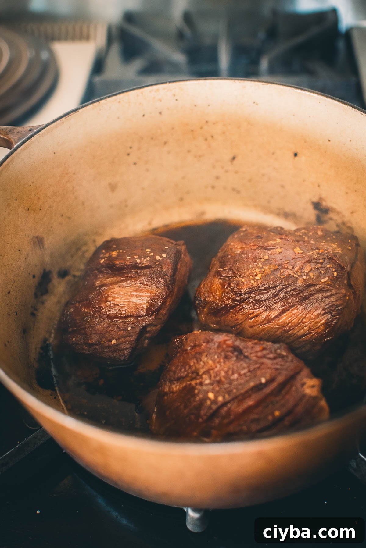Three pieces of browned short ribs cooking in a pot on a stovetop.