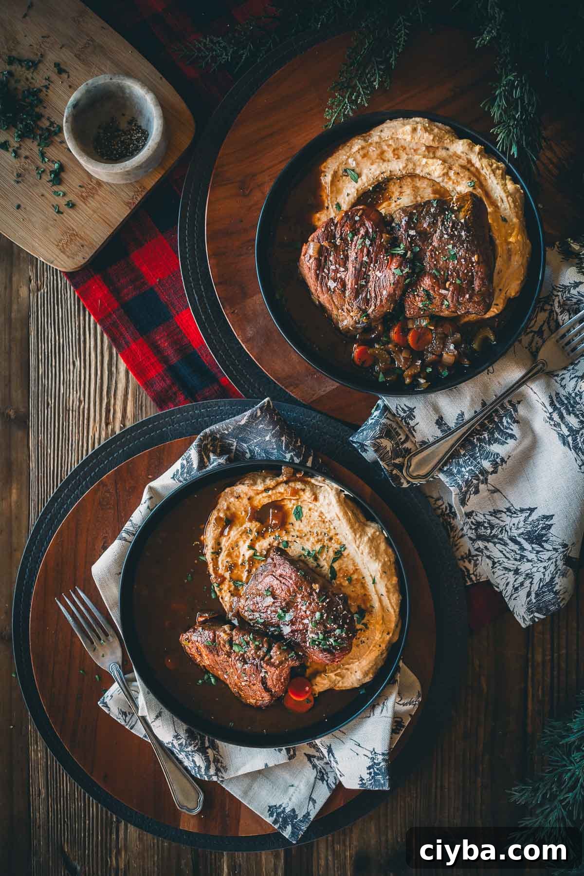 Two plates of sous vide short ribs with mashed root veggies and vegetables on a wooden table with decorative napkins and a plaid cloth.
