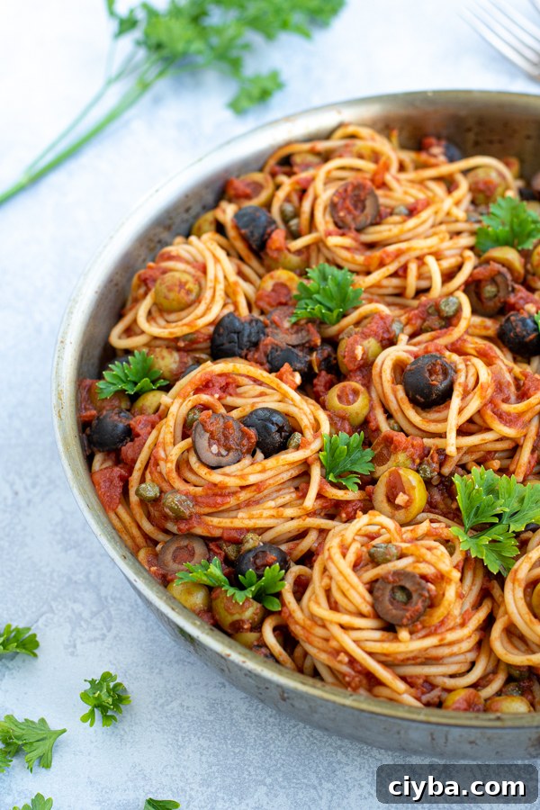 Authentic Spaghetti alla Puttanesca Recipe 6 close up of pasta twirled into nests inside of a large skillet garnished by olives and parsley