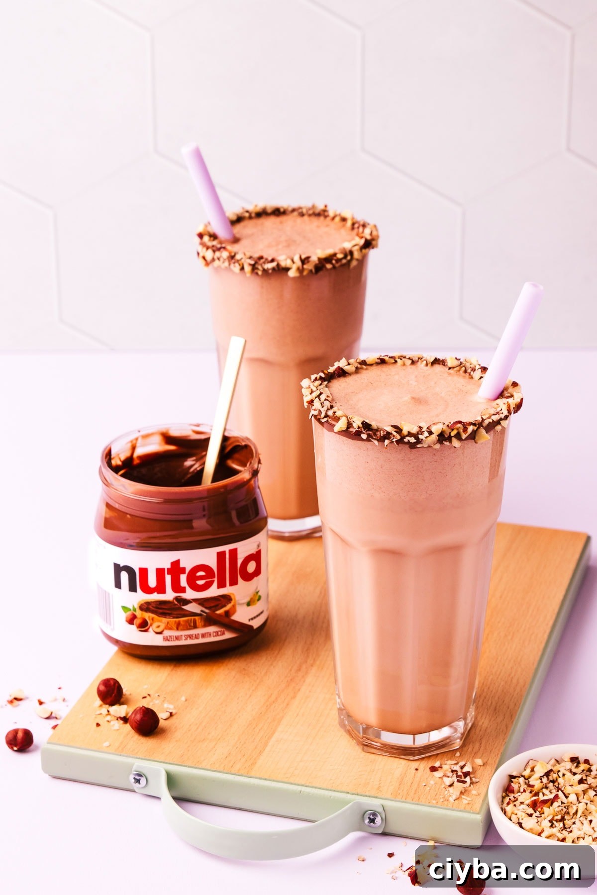 Two Nutella milkshakes on a small wooden chopping board, with an open jar of Nutella and scattered chopped and whole hazelnuts.