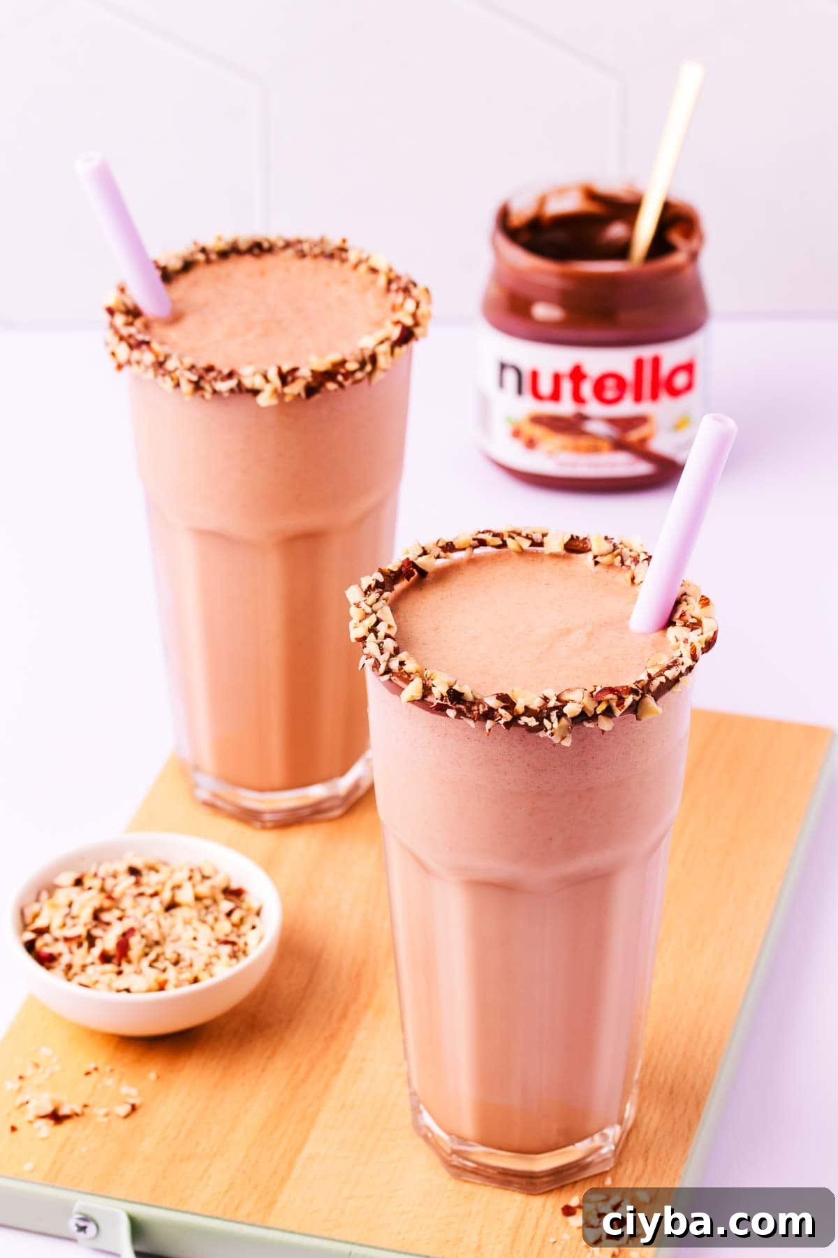 Two milkshakes and a small bowl of chopped hazelnuts on a small wooden board.