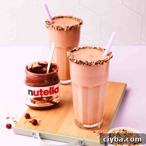 Two Nutella milkshakes on a small wooden chopping board, with an open jar of Nutella and scattered chopped and whole hazelnuts.