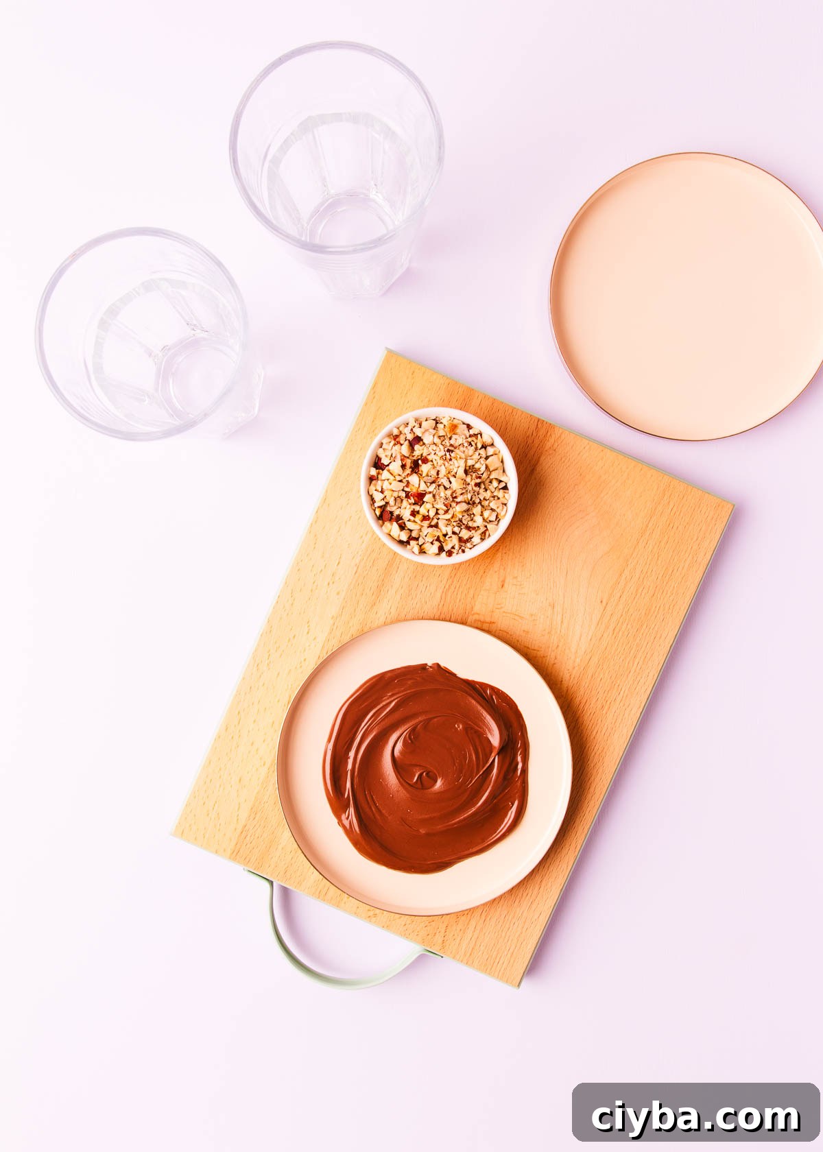 Softened Nutella spread onto a flat pink plate, with two tall empty glasses, on a pale purple background.