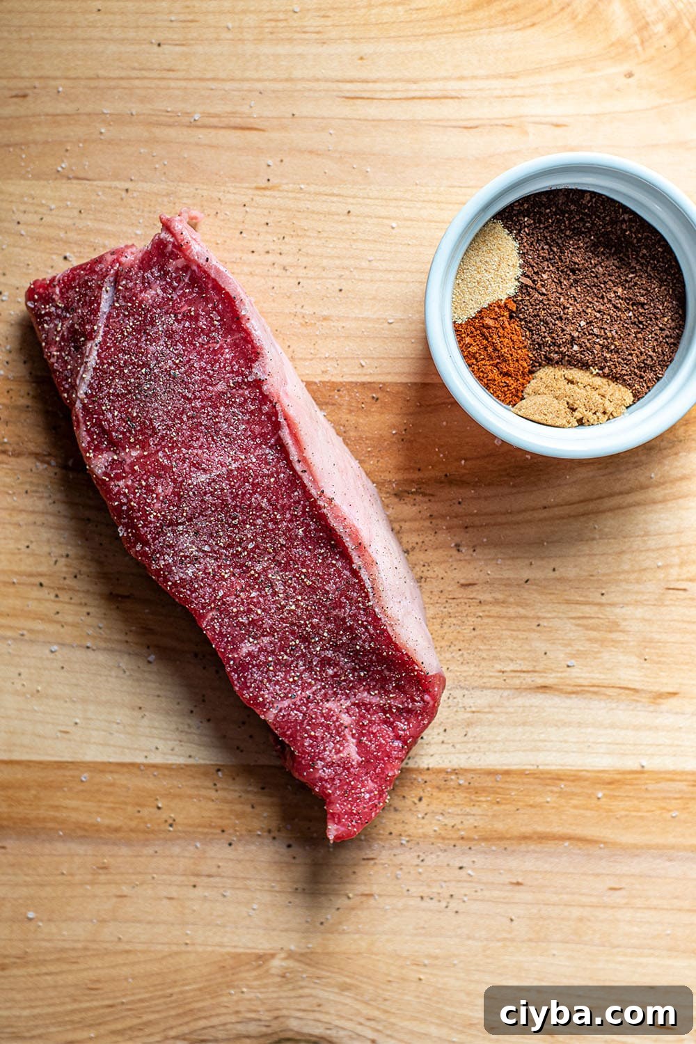 Coffee-Rubbed Steak Recipe for Tender, Flavorful Results 3 An uncooked seasoned NY strip steak next to a small white container filled with ground coffee and spices.