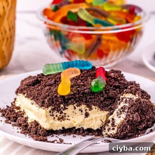 A dessert with a crumbly Oreo Dirt Cake topping and gummy worms on top, served on a white plate with a spoon; a bowl of gummy worms is in the background.