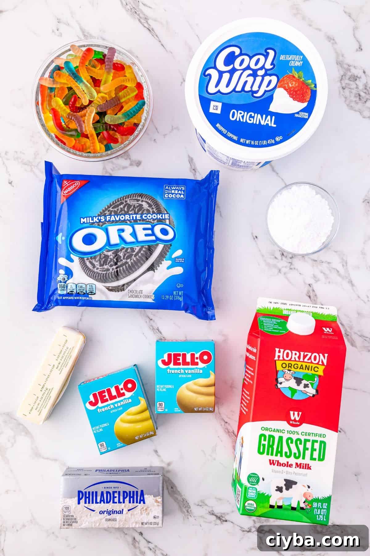 Ingredients on a marble counter gummy worms, Cool Whip, powdered sugar, Oreo cookies, butter, vanilla and chocolate Jell-O pudding, whole milk, and cream cheese.