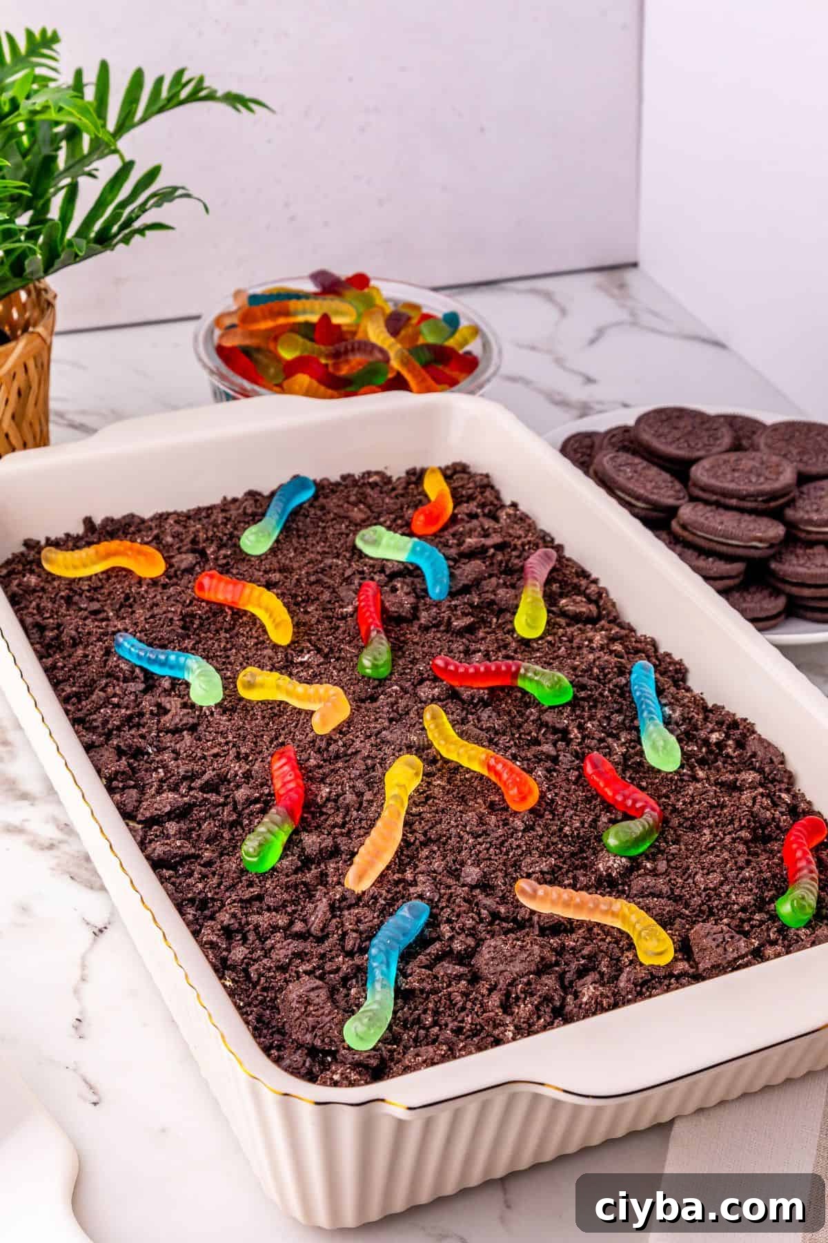 A rectangular dish filled with crumbled chocolate cookies and colorful gummy worms sits up front, with a bowl of gummy worms and a stack of sandwich cookies in the background.